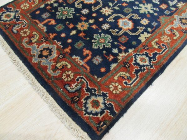 Ornate navy and rust rug corner with floral patterns and visible fringe on light blonde wood planks.