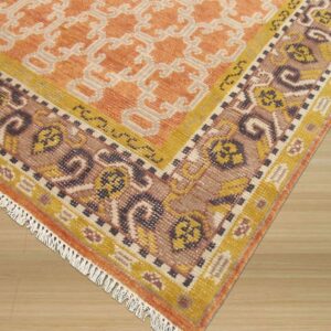 Orange, cream, and brown patterned rug with fringe and ornate borders shown on light wood flooring.