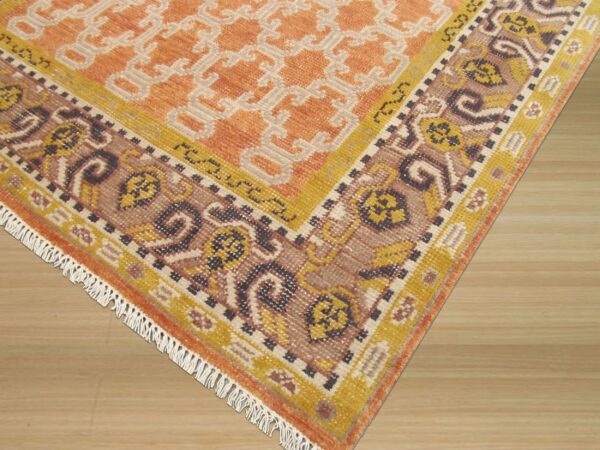 Orange, cream, and brown patterned rug with fringe and ornate borders shown on light wood flooring.