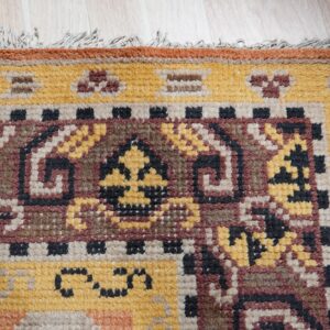 Textured rug corner displaying detailed geometric patterns in yellow, maroon, and dark brown on light wood.