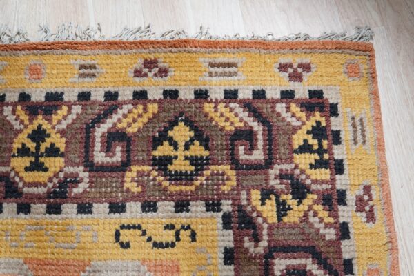 Textured rug corner displaying detailed geometric patterns in yellow, maroon, and dark brown on light wood.
