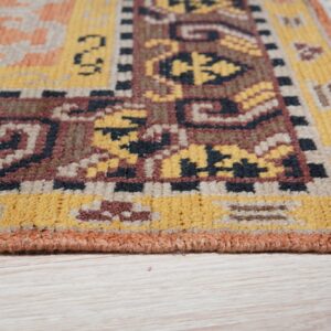The edge and corner of a patterned rug showing yellow, brown, and terracotta colors on pale wood flooring.