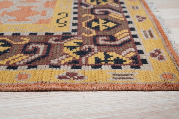 The edge and corner of a patterned rug showing yellow, brown, and terracotta colors on pale wood flooring.