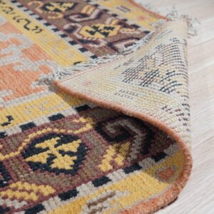 A geometric patterned rug in yellow, peach, and brown is rolled back on light plank flooring.
