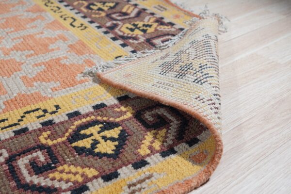 A geometric patterned rug in yellow, peach, and brown is rolled back on light plank flooring.