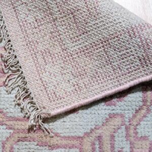 A fringed, high-pile geometric rug in pink and off-white is rolled back, showing the tightly woven underside.