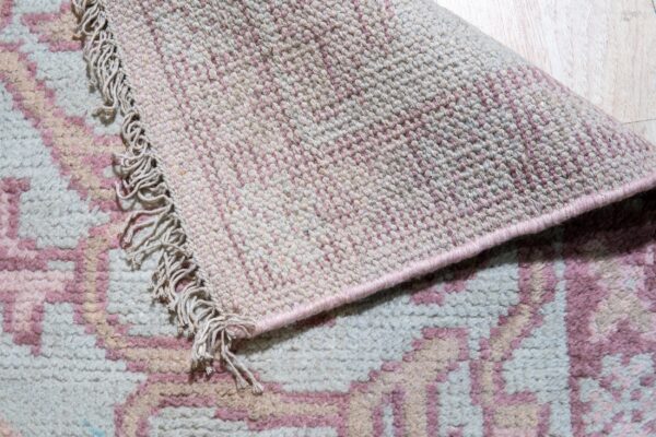 A fringed, high-pile geometric rug in pink and off-white is rolled back, showing the tightly woven underside.