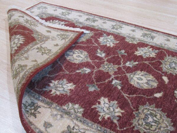 Close-up of a burgundy and beige floral rug folded back, resting on light wooden flooring.