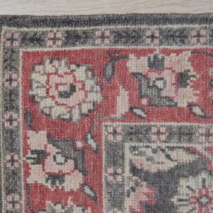 Textured rug corner in dusty red, gray, and cream with a traditional border pattern on light flooring.