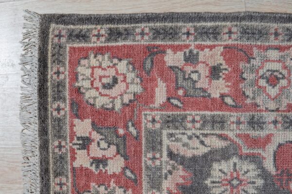 Textured rug corner in dusty red, gray, and cream with a traditional border pattern on light flooring.