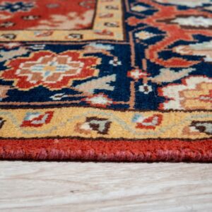 Traditional rug with navy field, gold border, and red serged edging sitting on pale wood flooring.