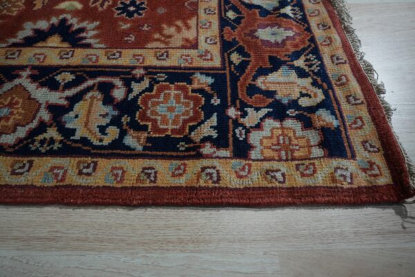 Medium-pile rug corner showing traditional navy, rust, and yellow floral patterns on pale wood floor.