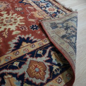 Rust and navy patterned rug with fringe is partially rolled back on light wood flooring.