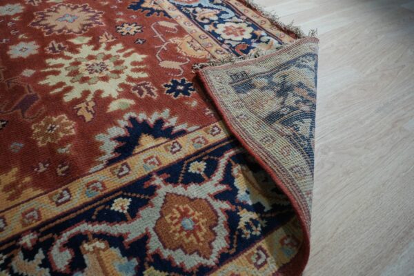 Rust and navy patterned rug with fringe is partially rolled back on light wood flooring.