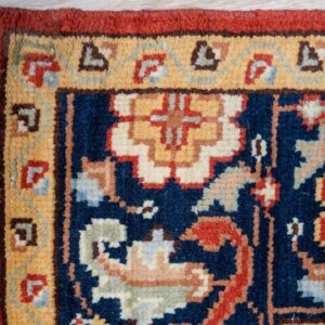 Navy rug corner with gold and red borders, intricate motifs, and fringe on light wood planks.