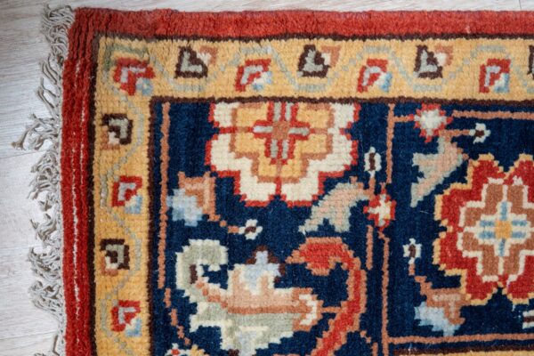 Navy rug corner with gold and red borders, intricate motifs, and fringe on light wood planks.