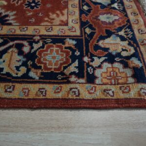 Ornate rug border in rust red, navy blue, and yellow with fringe on light wood planks.