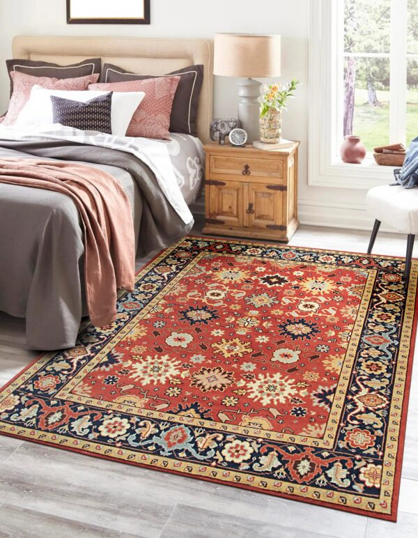 A rust-red traditional floral rug with navy borders lays on light wood flooring in a bright bedroom.