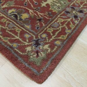 Corner detail of a textured, deep red rug with traditional multi-color floral patterns on pale wood floor.