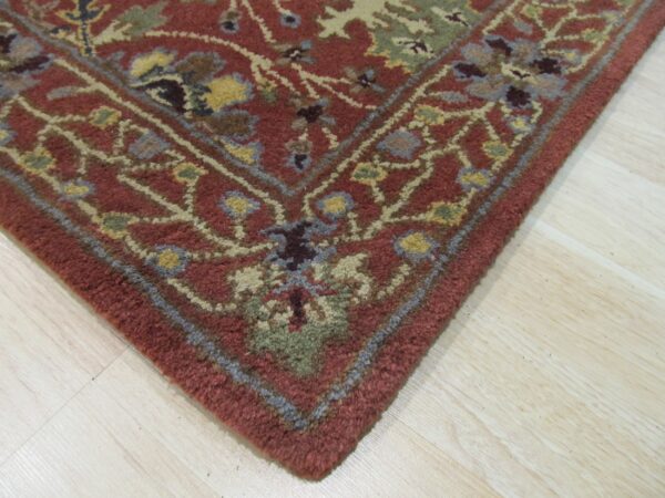 Corner detail of a textured, deep red rug with traditional multi-color floral patterns on pale wood floor.