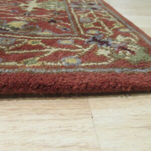 Thick-piled rug edge in rust red with multicolor traditional patterns on light wood flooring.