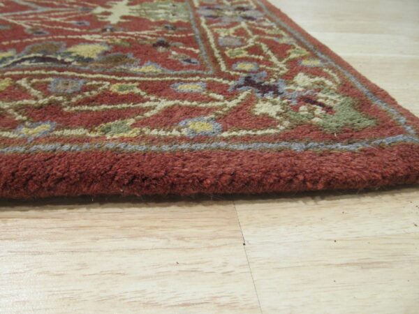 Thick-piled rug edge in rust red with multicolor traditional patterns on light wood flooring.