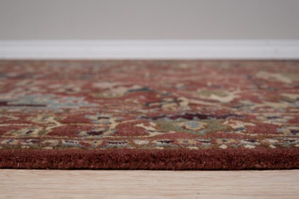 High-pile traditional rug border in reddish-brown resting on light wood floors against a gray wall.