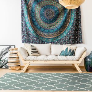A low-pile teal rug with a white quatrefoil pattern is beneath a light wood sofa and woven pendant lights.