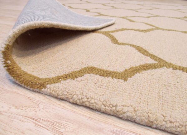 High-pile beige rug with olive trellis pattern curled up, showing its backing on blonde wood.