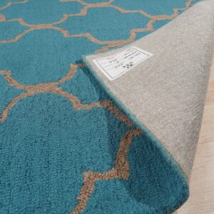 Teal rug with a tan geometric pattern is folded back, showing the backing and tag on light wood floors.