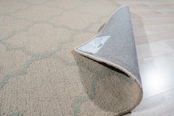 Textured beige and pale green rug shown curled back revealing the gray backing on light wood flooring.
