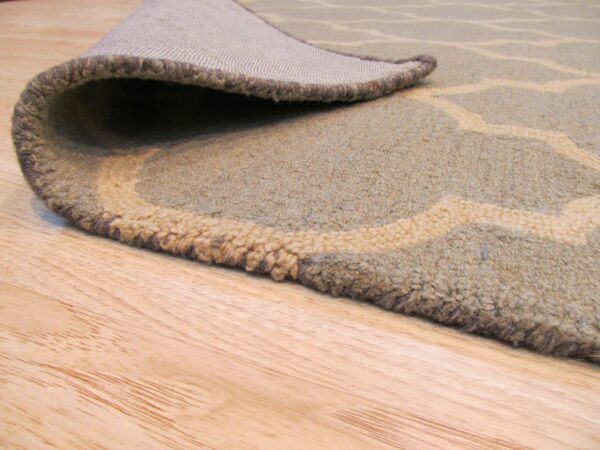 Muted green and beige patterned rug with visible pile and woven backing rests on wooden floor.