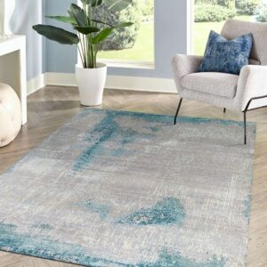A gray and teal abstract rug sits on wood chevron flooring in a sunlit room with a gray chair and large plant.