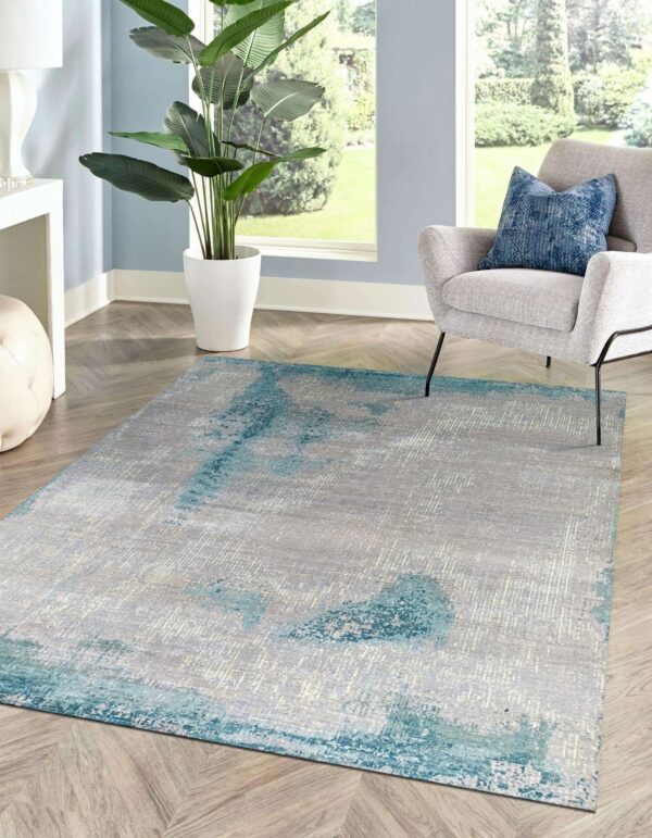 A gray and teal abstract rug sits on wood chevron flooring in a sunlit room with a gray chair and large plant.