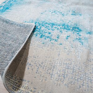 A gray and bright blue abstract rug is rolled up, revealing its woven backing on light plank flooring.
