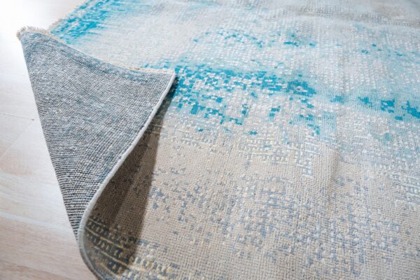 A gray and bright blue abstract rug is rolled up, revealing its woven backing on light plank flooring.