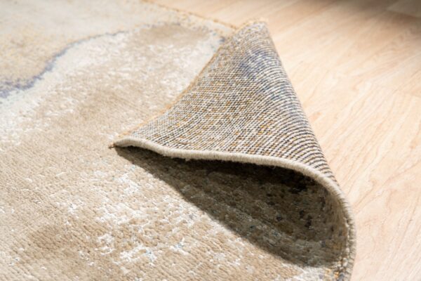 Medium-pile beige rug with abstract pattern folded to show the woven backing on light wood floors.
