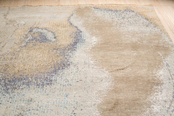 Close-up view of a beige rug featuring an abstract distressed pattern in gold, gray, and light blue.