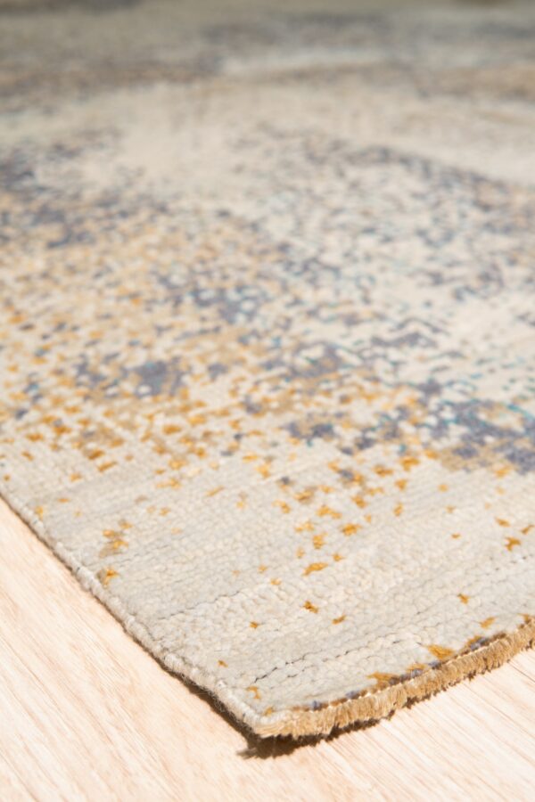 Textured light beige rug corner with scattered gold and grey pattern on light wood planks.