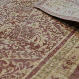 Beige rug with distressed maroon and green traditional patterns, edge curled back on light wood flooring.