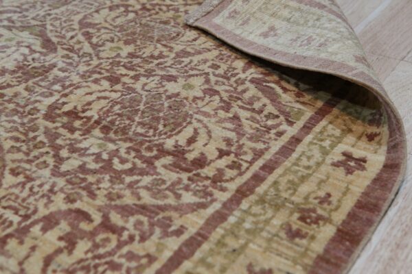 Beige rug with distressed maroon and green traditional patterns, edge curled back on light wood flooring.