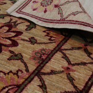 Close-up of a rolled tan rug featuring maroon and green stylized floral patterns on light wood flooring.