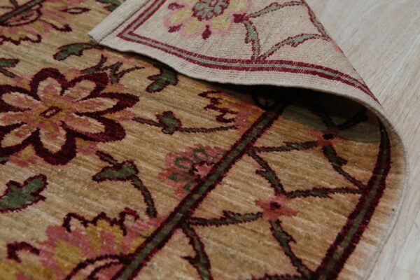 Close-up of a rolled tan rug featuring maroon and green stylized floral patterns on light wood flooring.