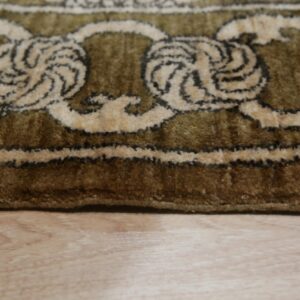 Olive green high-pile rug with beige and dark brown ornate scroll pattern on light wood floor.