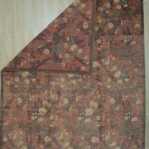 Rust-colored rug with dark scrolling floral patterns and cream flowers, partially folded on light wood floor.