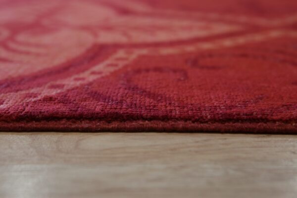 A deep red low pile rug edge rests on light wood planks, showing a subtle tonal pattern.