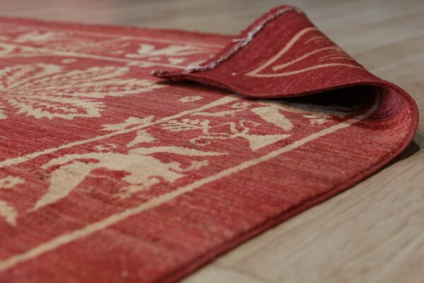 A close-up of a red and beige patterned flatweave rug curled on light wood floor.