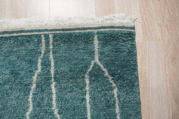 Textured teal rug with off-white geometric lines and fringe rests on light blonde wood planks.