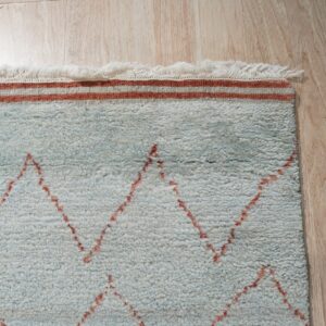 Pale blue shag rug with reddish-orange zigzag patterns and fringe rests on light wood flooring.