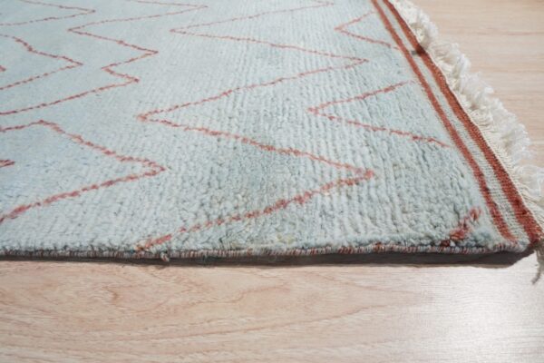 Mottled high-pile rug in light blue with red zigzag lines, showing fringe and light wood flooring.